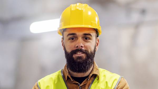 Young smiling construction worker in working clothes standing at construction site with arms crossed and looking at camera.